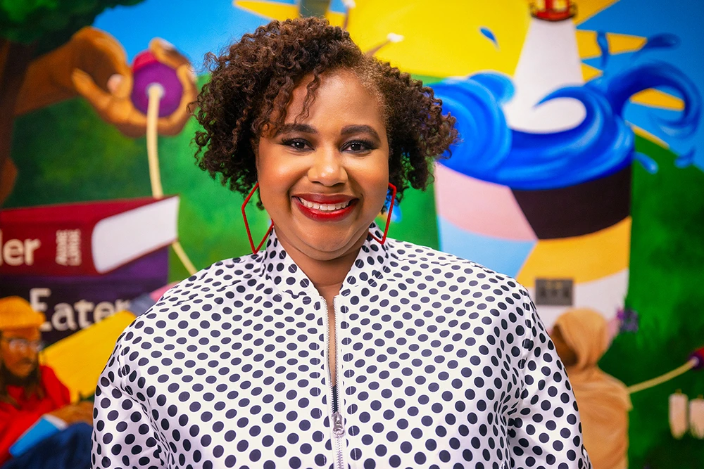 A woman wearing a black and white polka dot patterned shirt stands in front of a colorful mural.