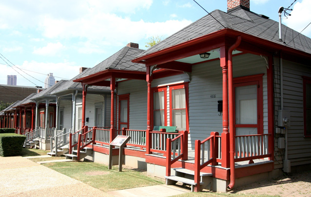 A row of single story houses with brightly painted front porch railings and small grass lots. A row of single story houses with brightly painted front porch railings and small grass lots.