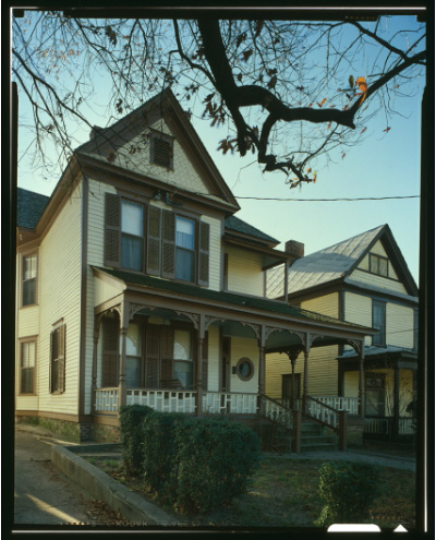 A narrow, two-story house with shutters, a porch, and low shrubs in front. A narrow, two-story house with shutters, a porch, and low shrubs in front.