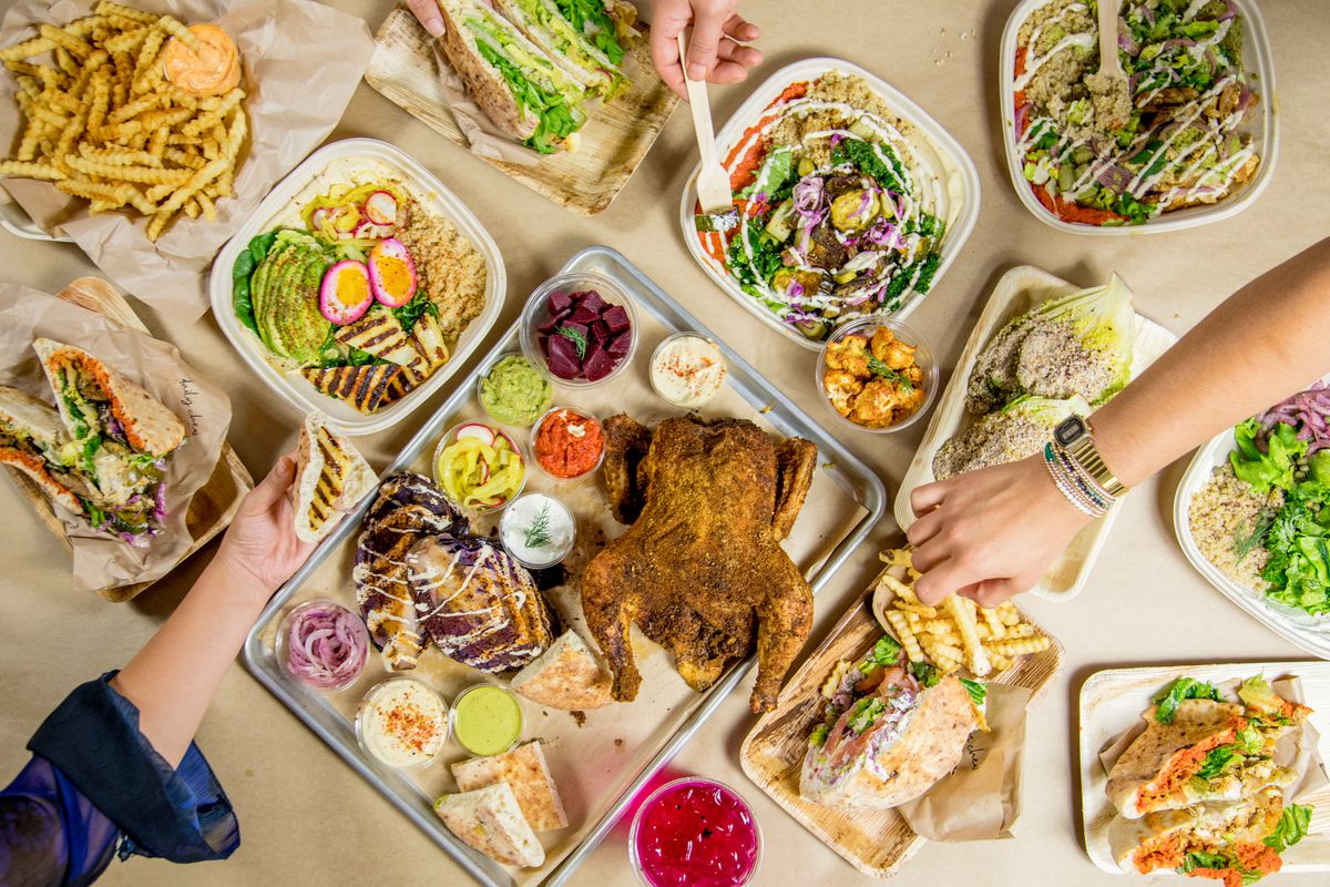 Hands reach out to sample a takeout spread of hummus, pita, a whole seasoned rotisserie chicken, chicken salad, pita with za’taar, and fries.