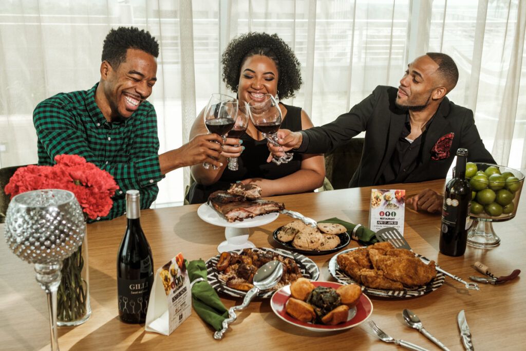 Black restaurant week founders Derek Robinson, Falayn Ferrell, and Warren Luckett laugh and cheers each other with wine at table full of food