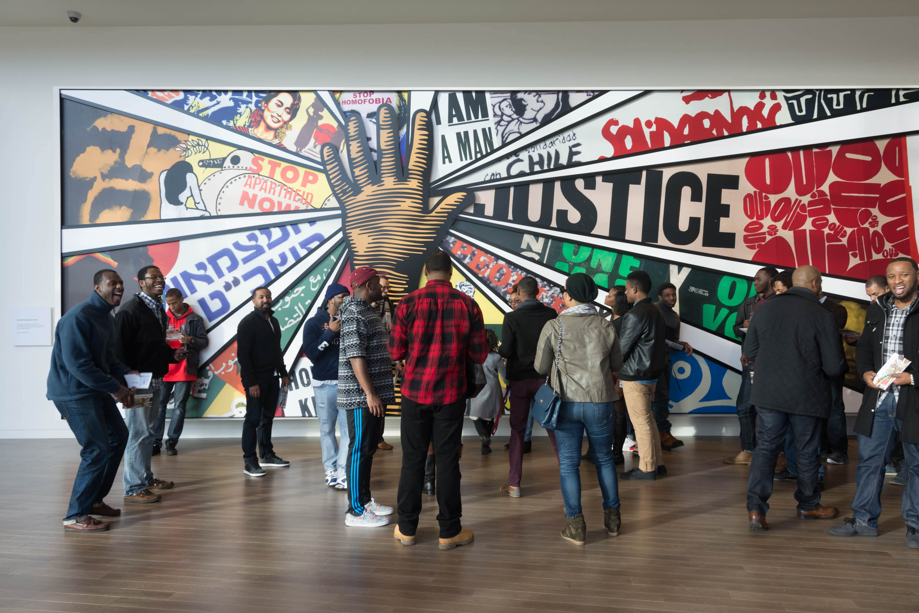 A group of people stand in front of a large painting with a black hand in the center and the word "justice" nearby