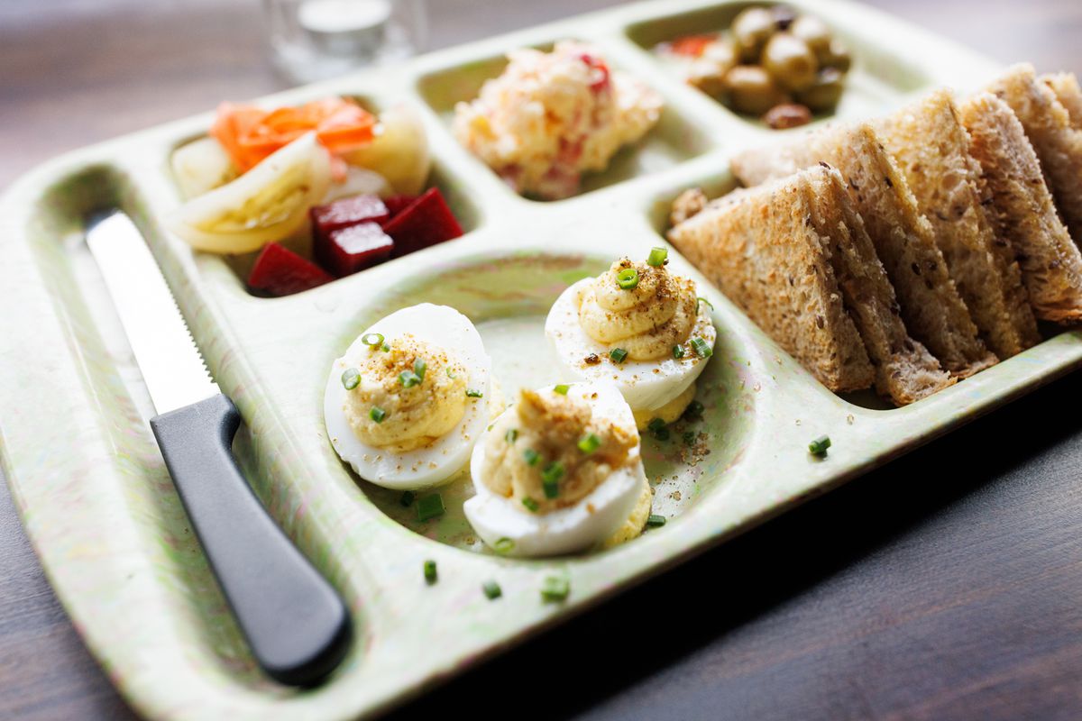 The Southern snack tray with pickled vegetables, pimento cheese, olives, deviled eggs, and toast points from Whoopsie’s in Atlanta sits in a plastic partitioned cafeteria tray.
